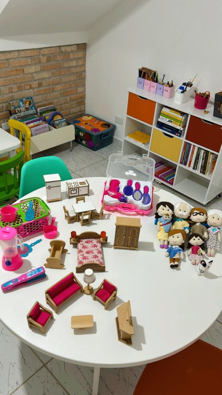 Mesa com materiais lúdicos: casinha de bonecas em madeira, família de bonecos de pano e brinquedos