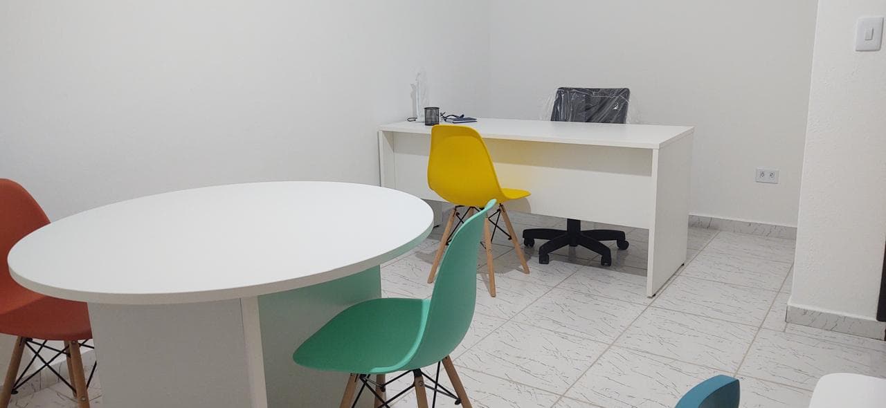Mesa redonda branca com cadeiras para a conversa com pais e adultos, e a mesa de trabalho ao fundo