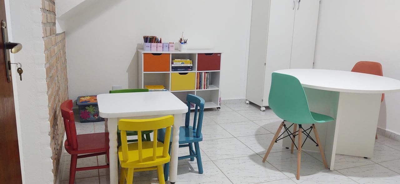 Sala de atendimento com mesa infantil e cadeiras coloridas, estante de materiais e mesa redonda de conversa